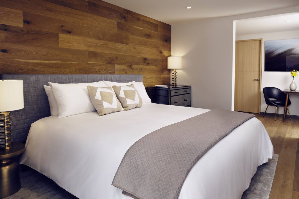 A modern hotel room with a large bed, a wooden headboard, a nightstand with a lamp, a desk with a chair, and a painting on the wall.