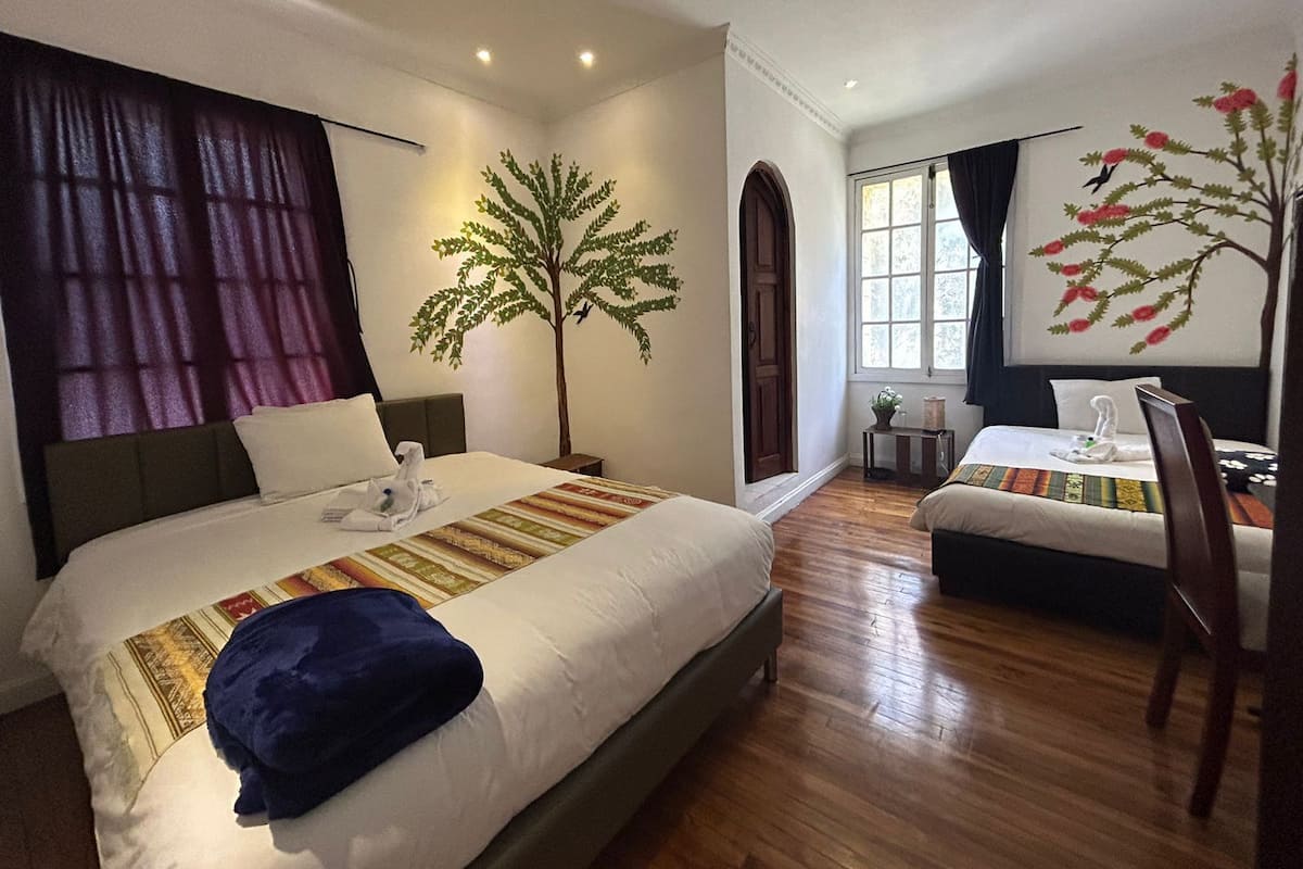 Une chambre d’hôtel avec un grand lit, un lit plus petit, un bureau et une chaise. Un mur est décoré d’une fresque représentant un arbre, et il y a une fenêtre avec des rideaux.