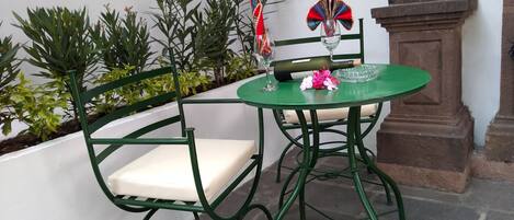 A green metal chair and table set on a balcony with a white wall and plants in the background.