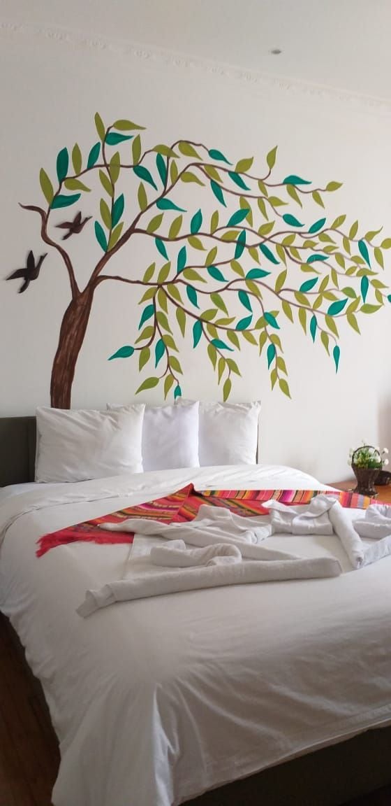 Una cama con ropa blanca, una colcha colorida y un mural en la pared con un árbol de hojas verdes y dos pájaros.
