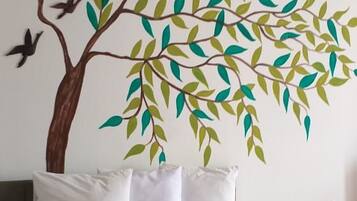 Una cama con ropa blanca, una colcha colorida y un mural en la pared con un árbol de hojas verdes y dos pájaros.