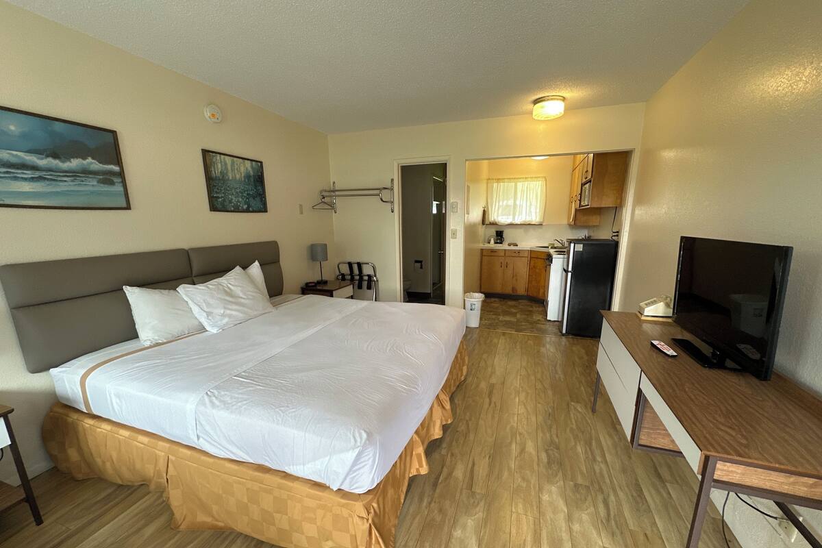 A hotel room with a large bed, a desk, a television, a kitchen area, and a bathroom visible through an open door.