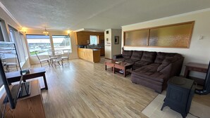 Amplia sala de estar con un sofá marrón grande, una mesa de centro de madera y un comedor con sillas. Se observa una cocina al fondo, y hay un ventanal con vistas al exterior.