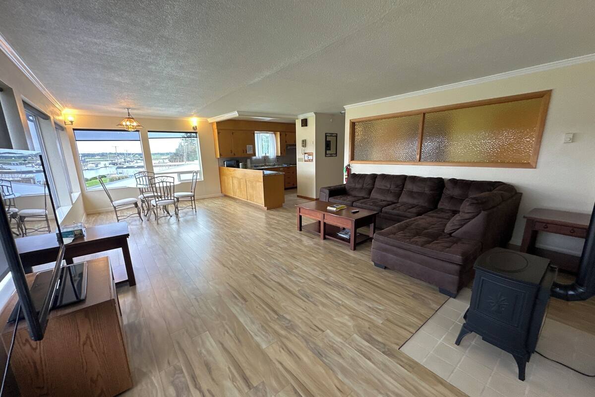 A spacious living area with a large brown sofa, a wooden coffee table, and a dining area with chairs. A kitchen is visible in the background, and there is a large window offering a view of the outdoors.
