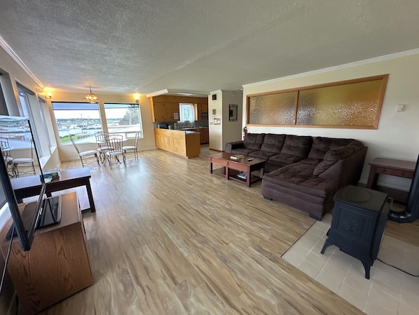 A spacious living area with a large brown sofa, a wooden coffee table, and a dining area with chairs. A kitchen is visible in the background, and there is a large window offering a view of the outdoors.