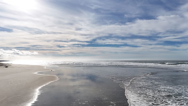 Una playa con una amplia orilla arenosa, olas suaves y cielo parcialmente nublado.