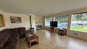 Una sala de estar con un sofá modular, una mesa de centro, un televisor y ventanas amplias que ofrecen vistas a un edificio y follaje.