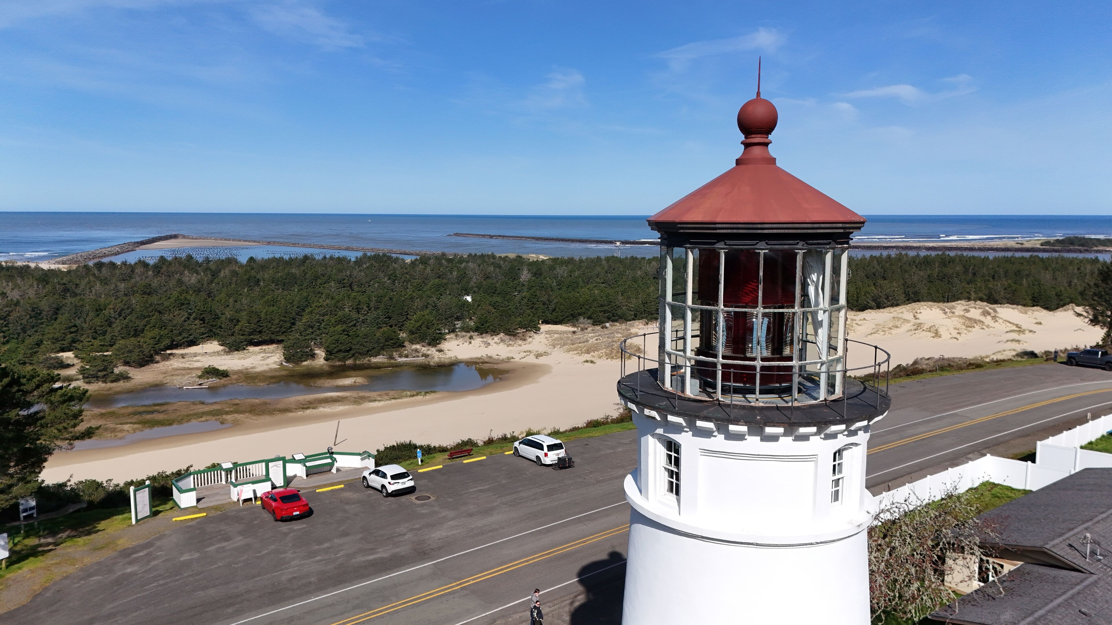 Un phare coiffé d’un dôme rouge, entouré d’un parking où sont garées plusieurs voitures, d’une plage de sable et, au loin, d’une zone boisée.