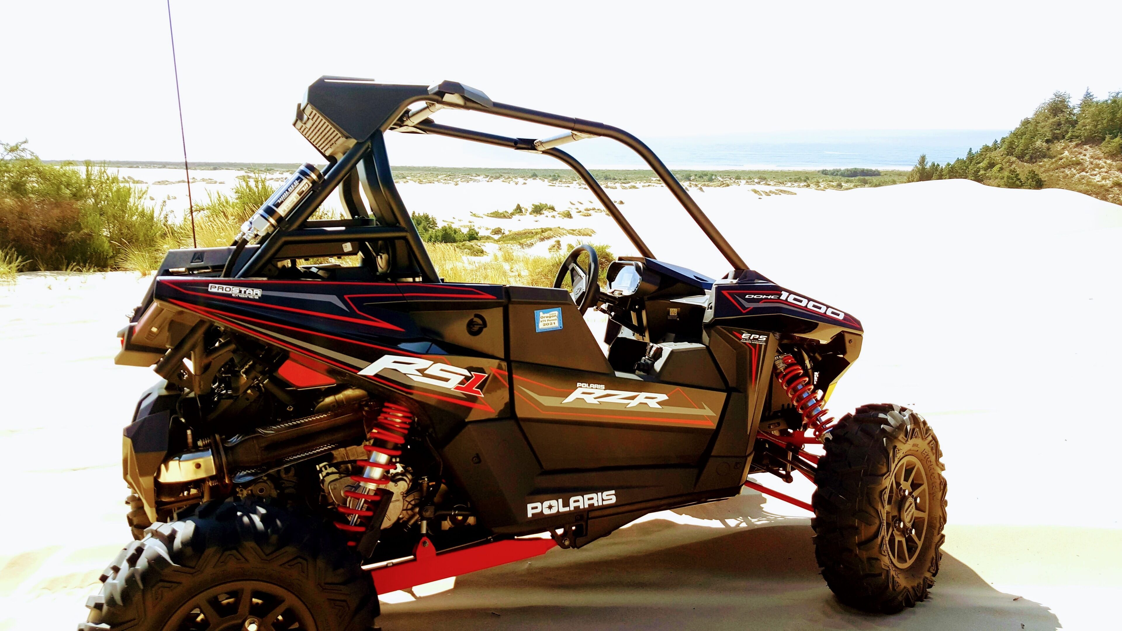 A Polaris RZR 1000 off-road vehicle parked on a sandy terrain with a clear sky in the background.