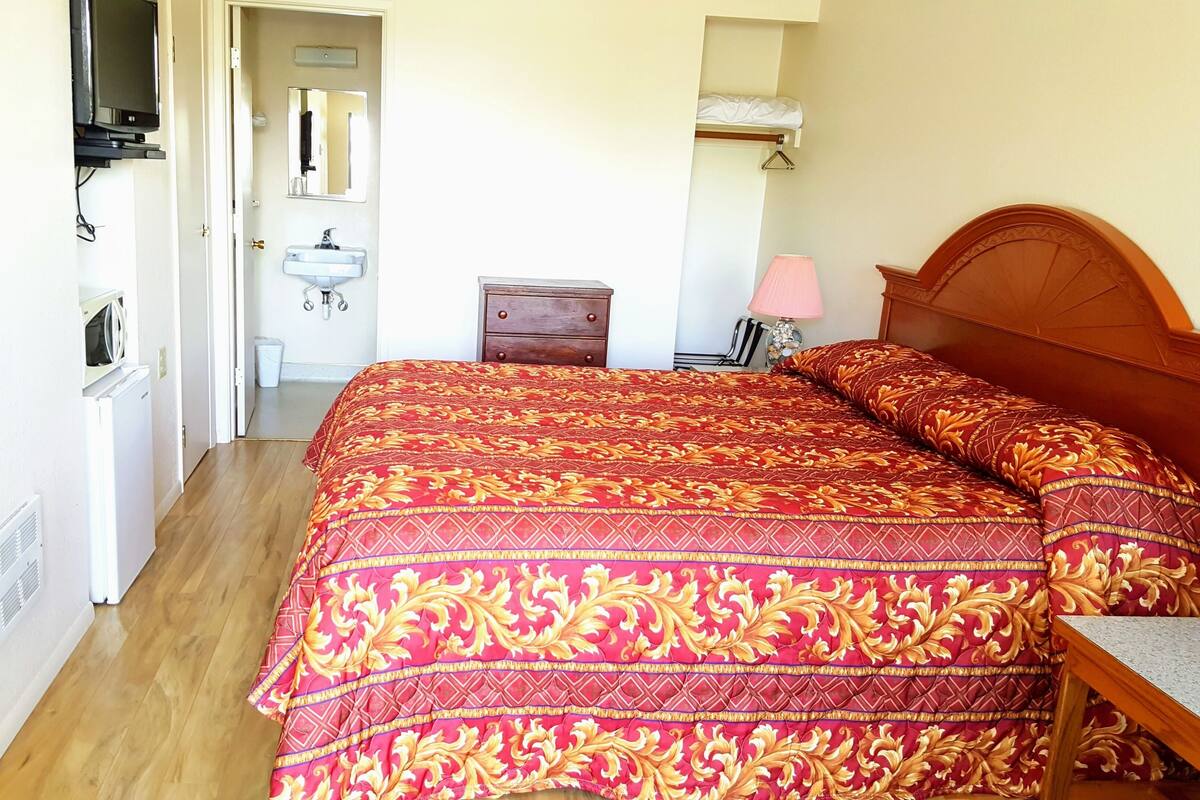 A bedroom with a large bed, a wooden nightstand, a bedside lamp, a dresser, and a television on a stand.