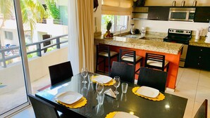 A dining area with a table set for four, a kitchen with modern appliances, and a view of the outdoors. - Condo Pacifico by Playa Paradise (Playa del Carmen)
