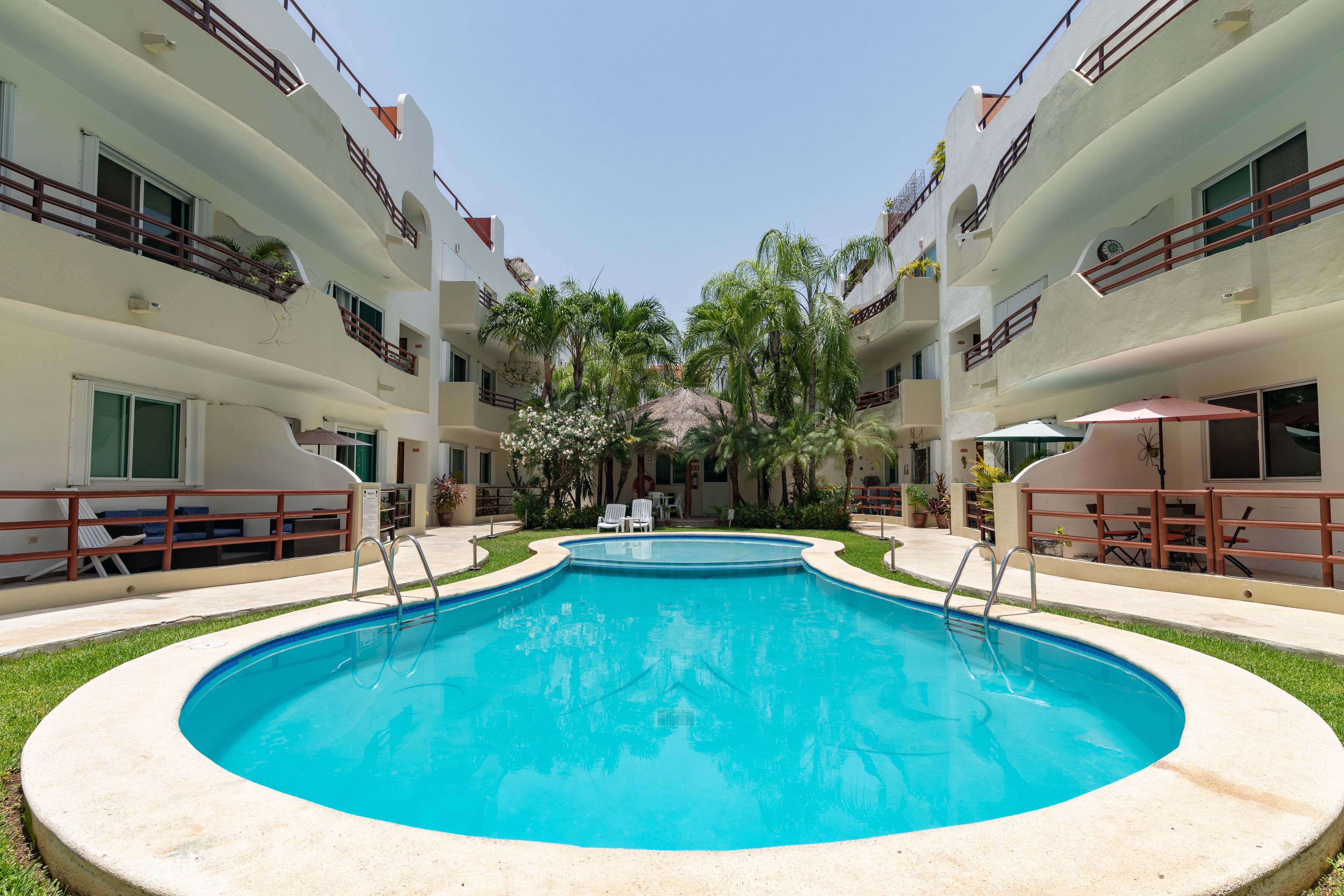 A swimming pool surrounded by a building with balconies, palm trees, and outdoor seating.