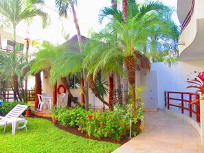 A courtyard with palm trees, a small garden with red flowers, and a white building with a clock on the wall. - Condo Margaritas By Playa Paradise (Playa del Carmen)