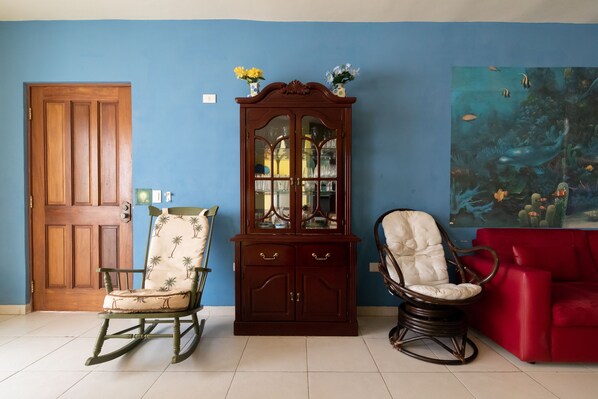 A room with a wooden door, a rocking chair with a palm tree pattern, a cabinet with glass doors, and a red sofa.