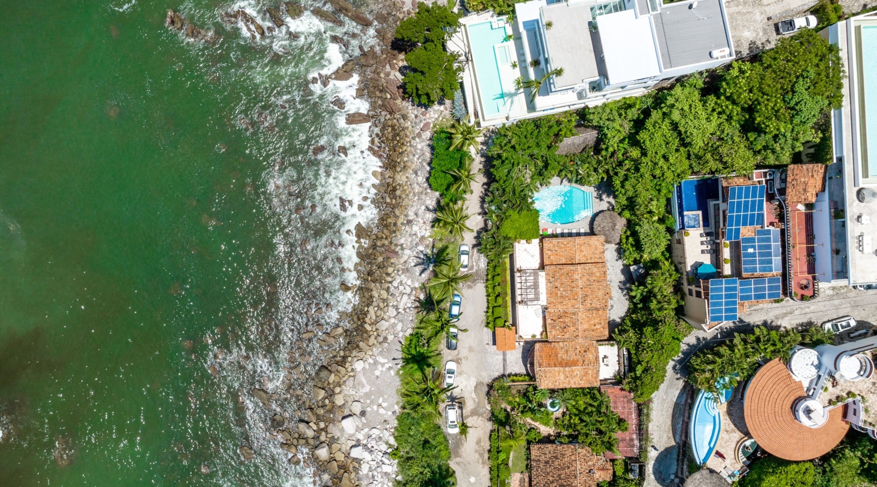An aerial view of a coastal property with a swimming pool, solar panels, and a beach area.