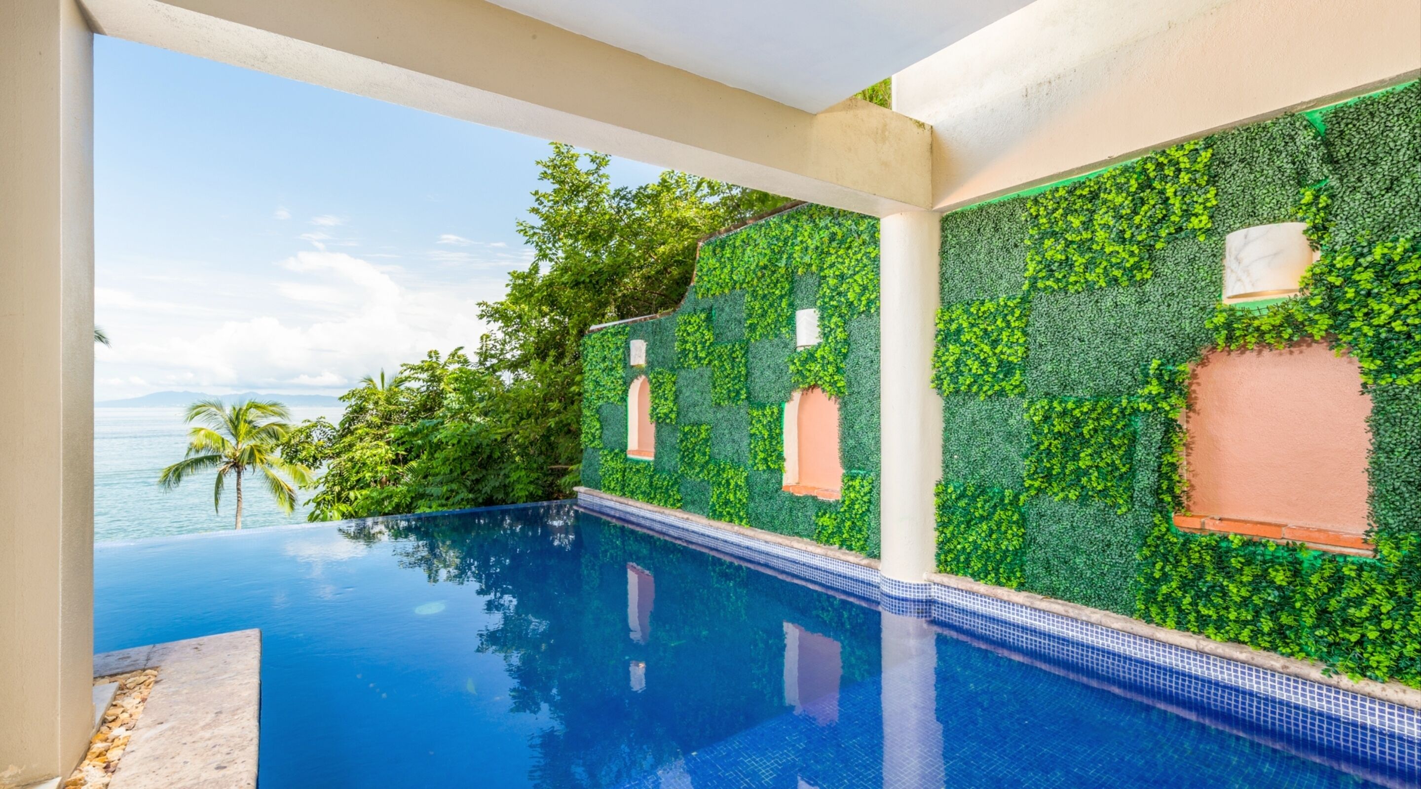 A pool with a clear view of the ocean and palm trees, surrounded by walls covered in green foliage.