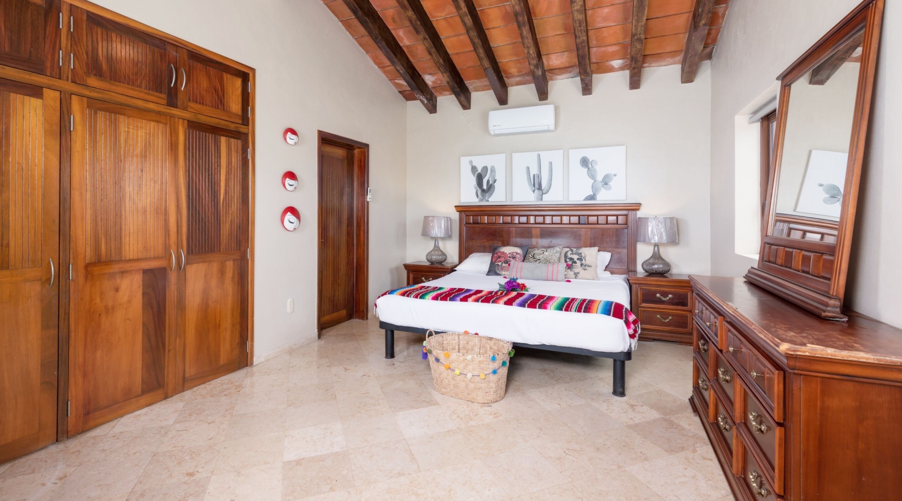 Un dormitorio con un armario grande de madera, una cama con manta colorida, dos mesitas de noche con lámparas, un espejo y un techo con vigas a la vista.