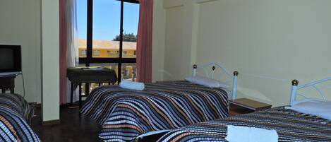 A hotel room with three beds, a television, and a window with a view of buildings outside.