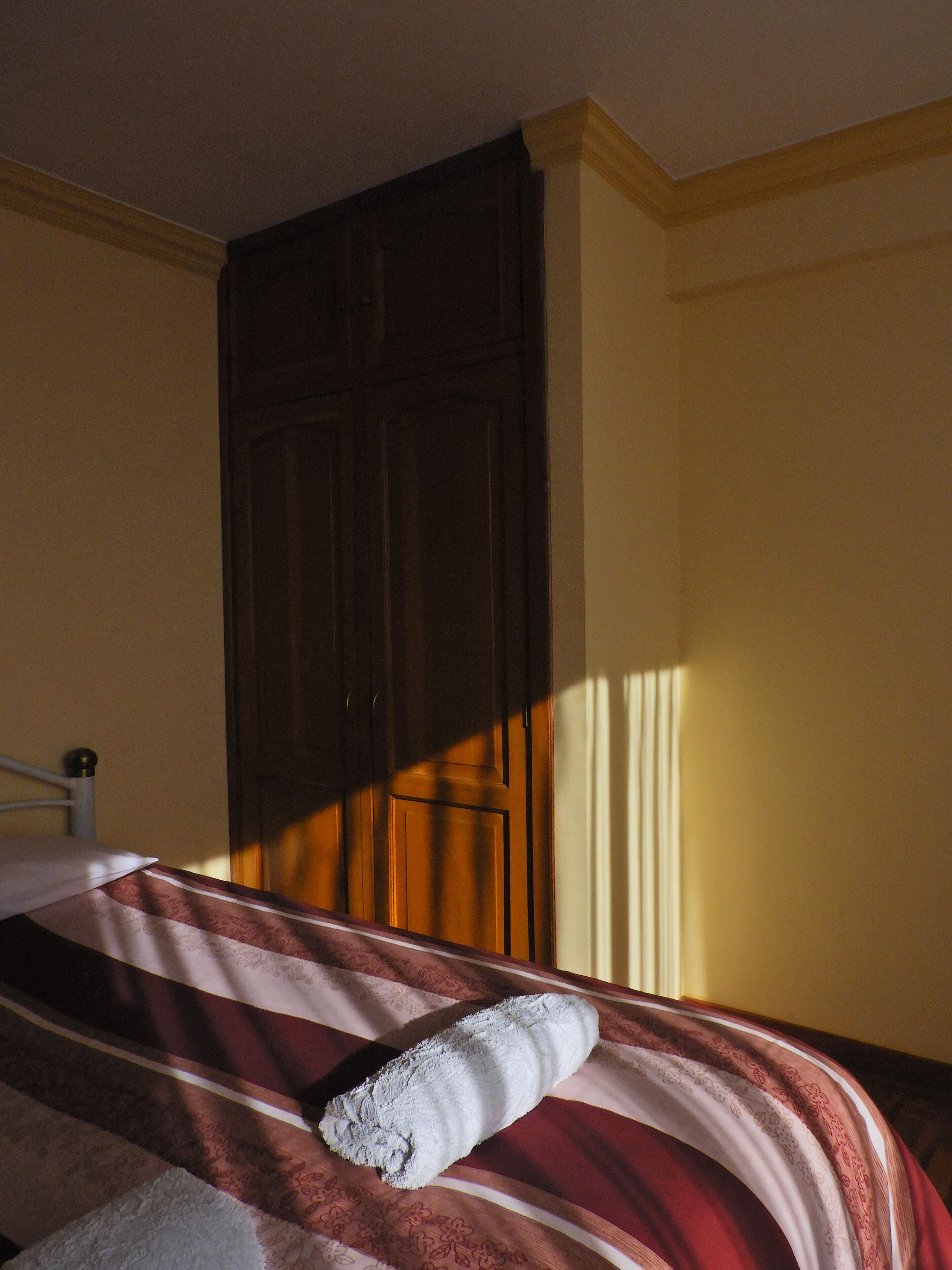 Una cama con una manta a rayas y una toalla enrollada, un armario de madera y una ventana que ilumina la pared.