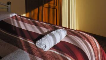 Una cama con una manta a rayas y una toalla enrollada, un armario de madera y una ventana que ilumina la pared.