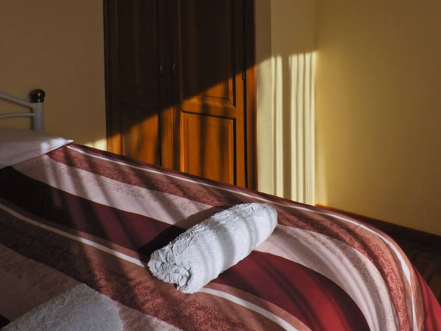 Una cama con una manta a rayas y una toalla enrollada, un armario de madera y una ventana que ilumina la pared.