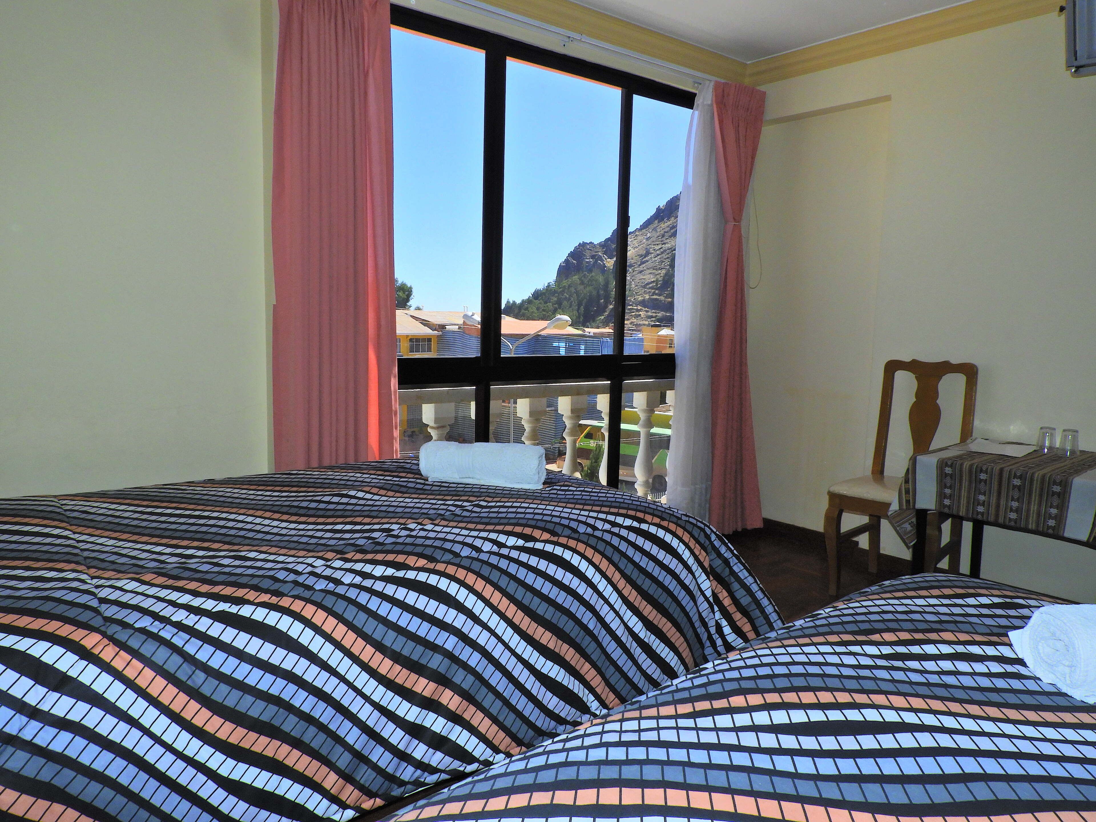 Habitación de hotel con dos camas, vistas a las montañas y un balcón con mesa y silla.