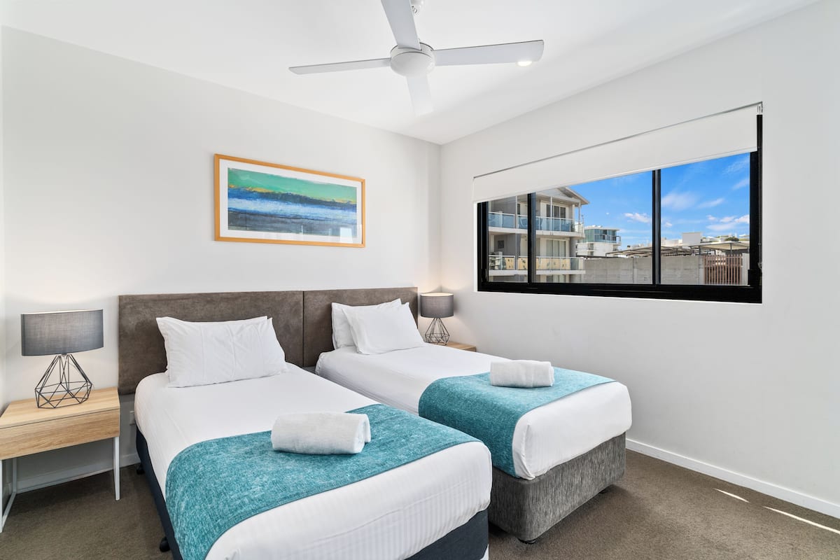 A hotel room with two beds, a ceiling fan, a painting, a window with a city view, and a nightstand with a lamp.