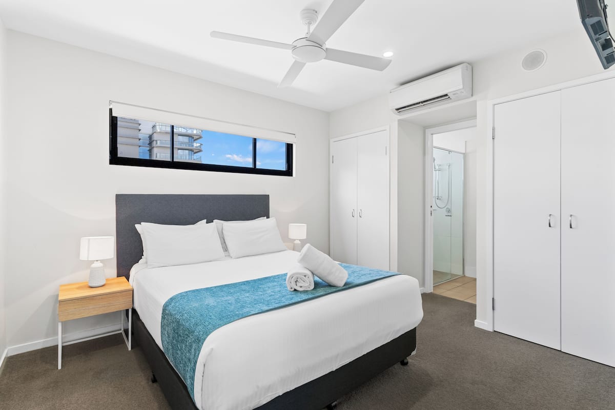 A modern hotel room with a bed, a ceiling fan, an air conditioning unit, a nightstand with a lamp, a window with a view of buildings, and a wardrobe.