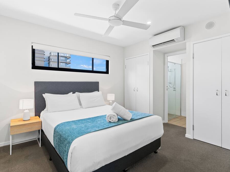 A modern hotel room with a bed, a ceiling fan, an air conditioning unit, a nightstand with a lamp, a window with a view of buildings, and a wardrobe.