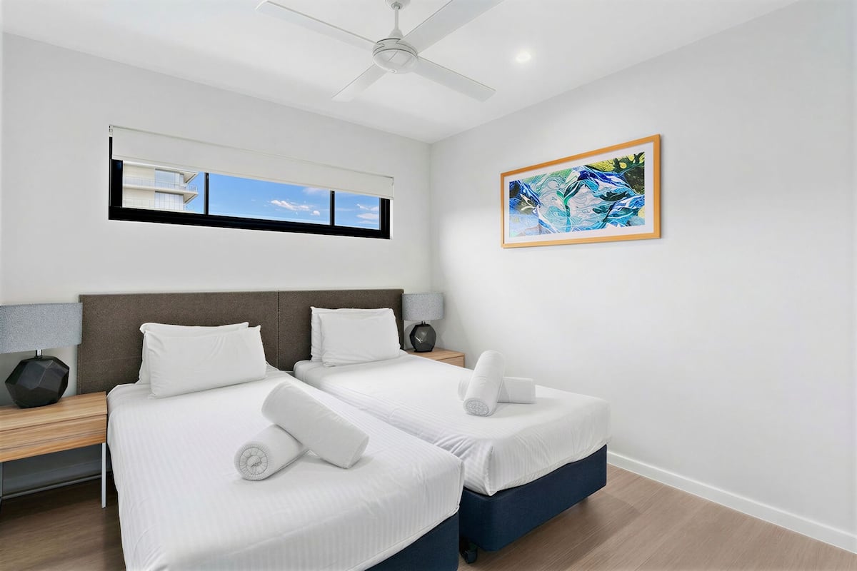 A hotel room with two beds, a ceiling fan, a window with a view, a framed artwork, and a nightstand with a lamp.