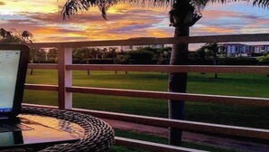 Una computadora portátil sobre una silla de mimbre, con vista a un atardecer y un área verde más allá de una cerca.