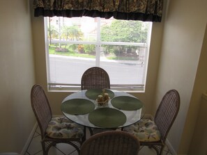 Un coin repas avec une table ronde, deux chaises en osier et une fenêtre avec des stores.