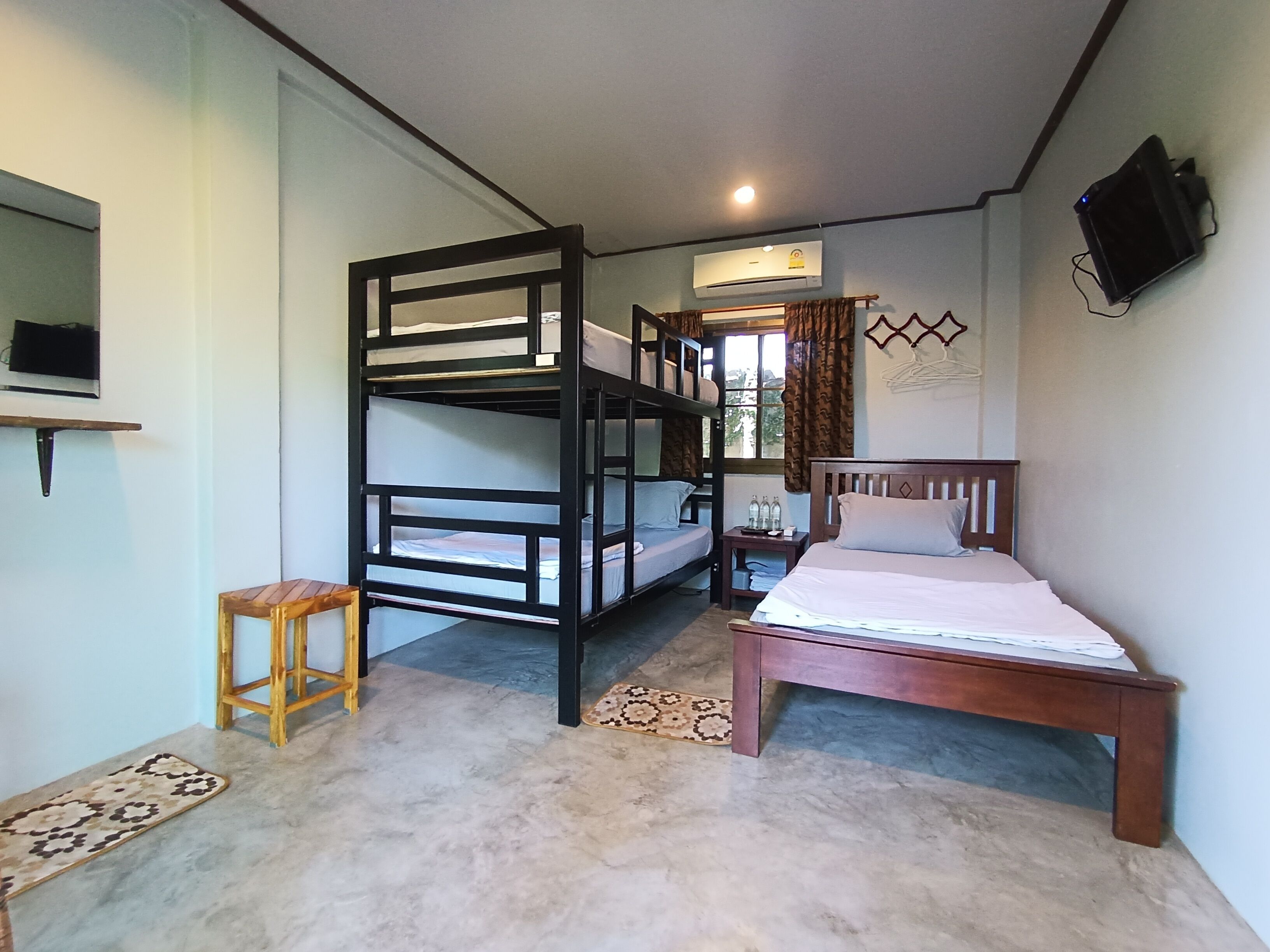 A bedroom with bunk beds, a single bed, a small wooden stool, a bedside table with bottles, and a mounted TV.
