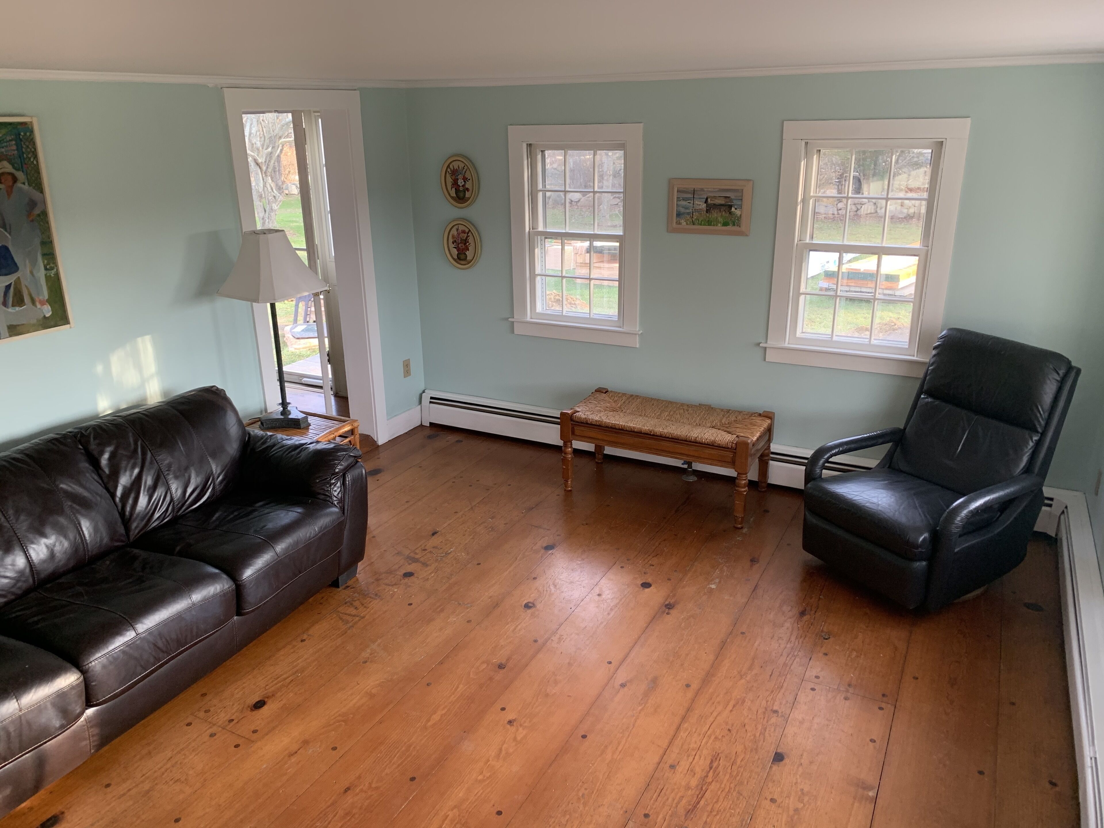 A living room with a black leather couch, a wooden bench, and a black leather chair. There are two windows with white frames and a door leading outside.