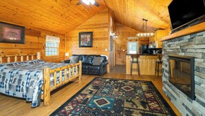 A log cabin interior with a bed, sofa, fireplace, and kitchen area.