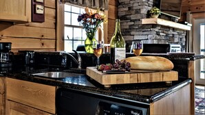 A kitchen with wooden cabinets, a stone wall, a wooden countertop, a dishwasher, and a cutting board with fruits and a bottle.