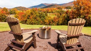 Two Adirondack chairs facing a metal fire pit with logs, set on a gravel area with a scenic view of autumn-colored trees and mountains in the background.