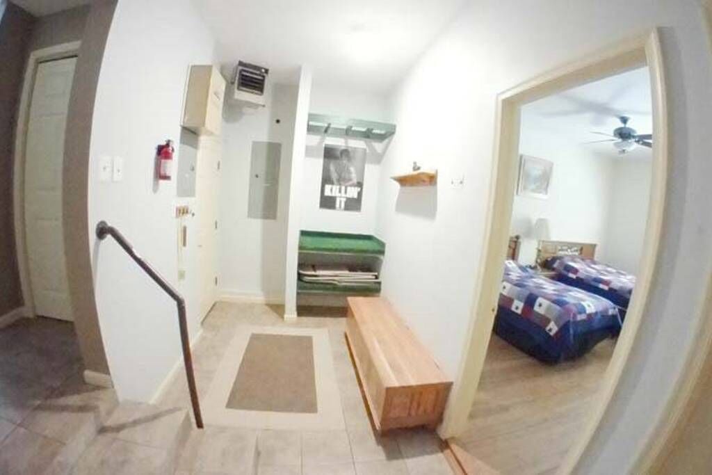 A narrow hallway with a wooden bench, a green mat, and a bed with a patterned blanket.