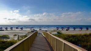 A wooden boardwalk leading to a beach with blue umbrellas and lounge chairs.