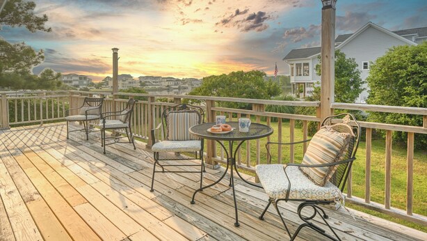 A wooden deck with outdoor furniture, a table with a plate of food, and a view of houses and trees in the background. - Breathtaking Sunsets On Back Bay- Your Sandbridge Escape Awaits! (Virginia Beach)