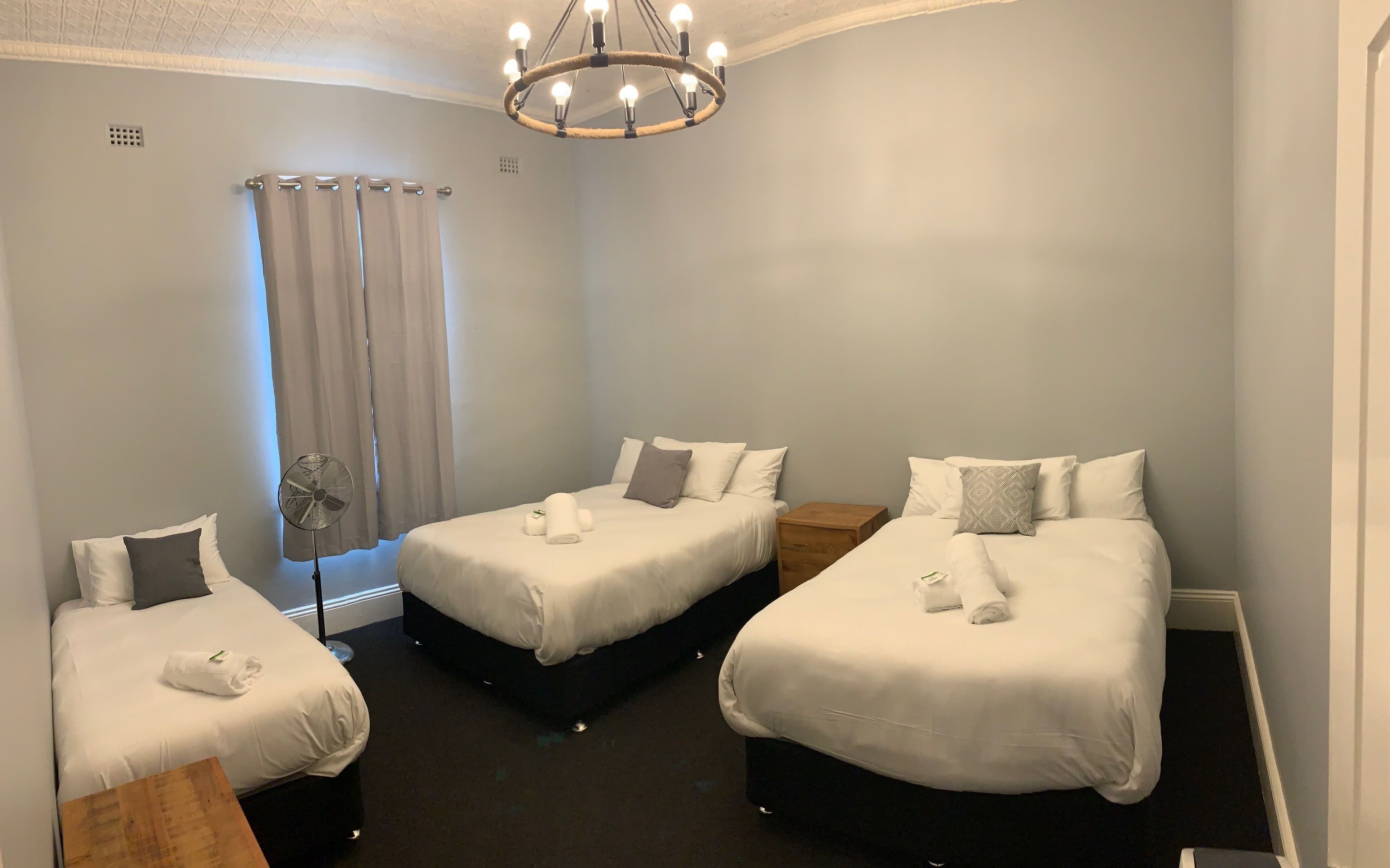 Quarto de hotel com três camas de solteiro, um ventilador de teto, uma mesa de cabeceira de madeira e um lustre.