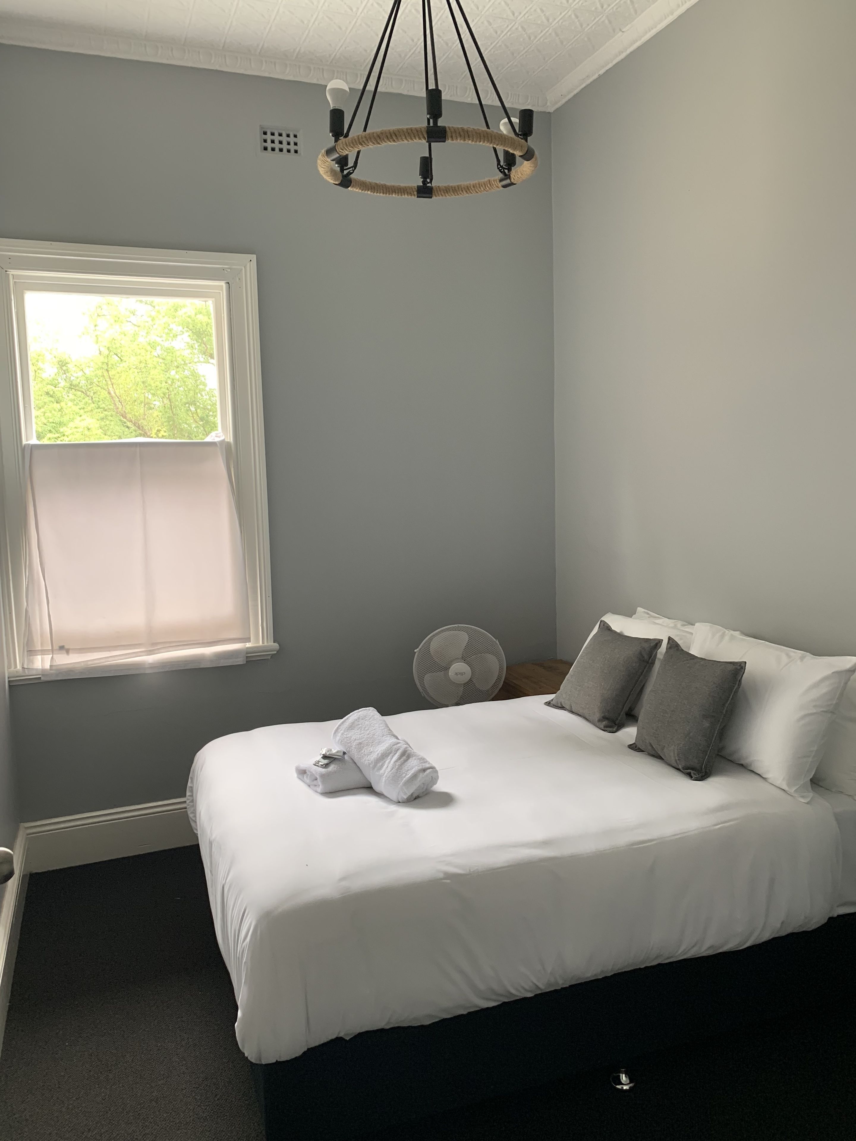 A neatly made bed with white bedding, a standing fan, and a window with sheer curtains.