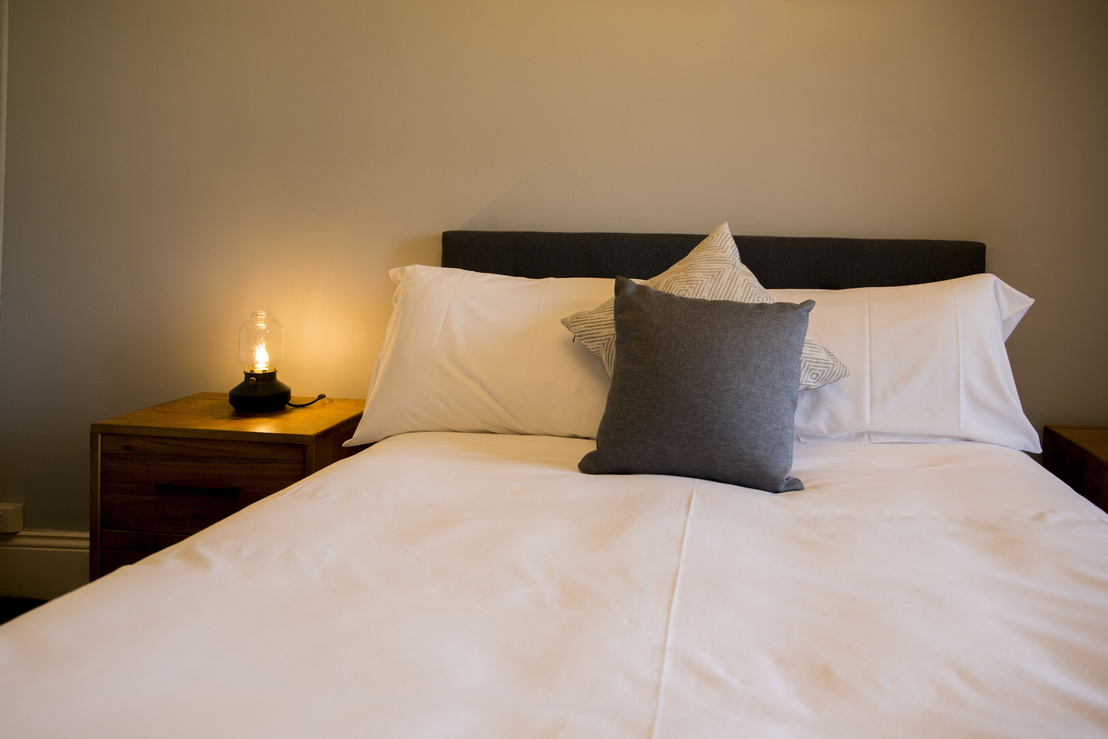 Una cama bien tendida con sábanas blancas y dos almohadas, una gris y otra a rayas, en una habitación sencilla con una mesita de noche de madera y una lámpara de mesa pequeña.