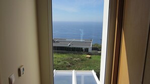 A view through a window of a building with a flat roof, a grassy area, and a body of water in the background. - CASA IVA 3 bedroom, 3 bathroom House with swimming pool and sea views (Estreito da Calheta)