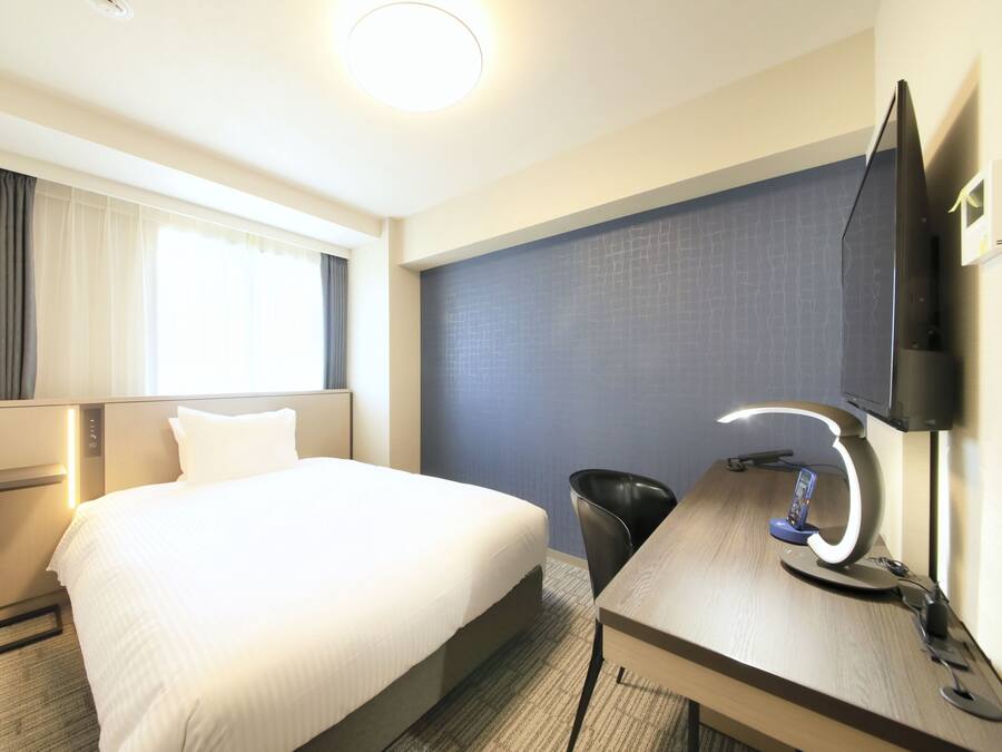 A hotel room with a large bed, a desk with a computer and a lamp, a TV mounted on the wall, and a window with curtains.