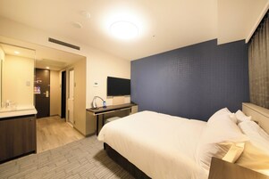 A hotel room with a bed, a desk, a chair, a flat-screen TV, and a bathroom with a mirror and sink.