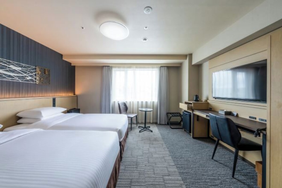 A hotel room with two beds, a desk, a chair, a TV, and a window with curtains.