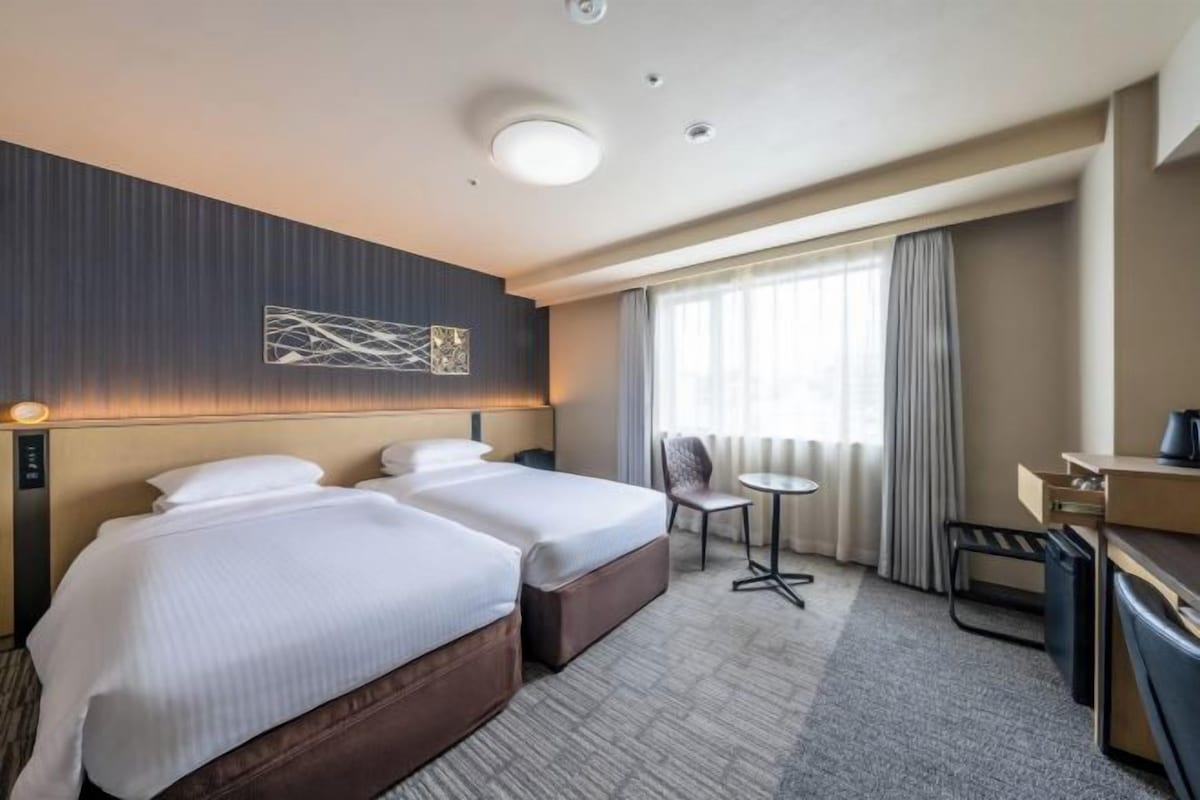 A hotel room with two beds, a desk, a chair, and a small table.