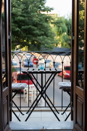 A balcony with a table and two chairs, overlooking a street with parked cars and trees.
