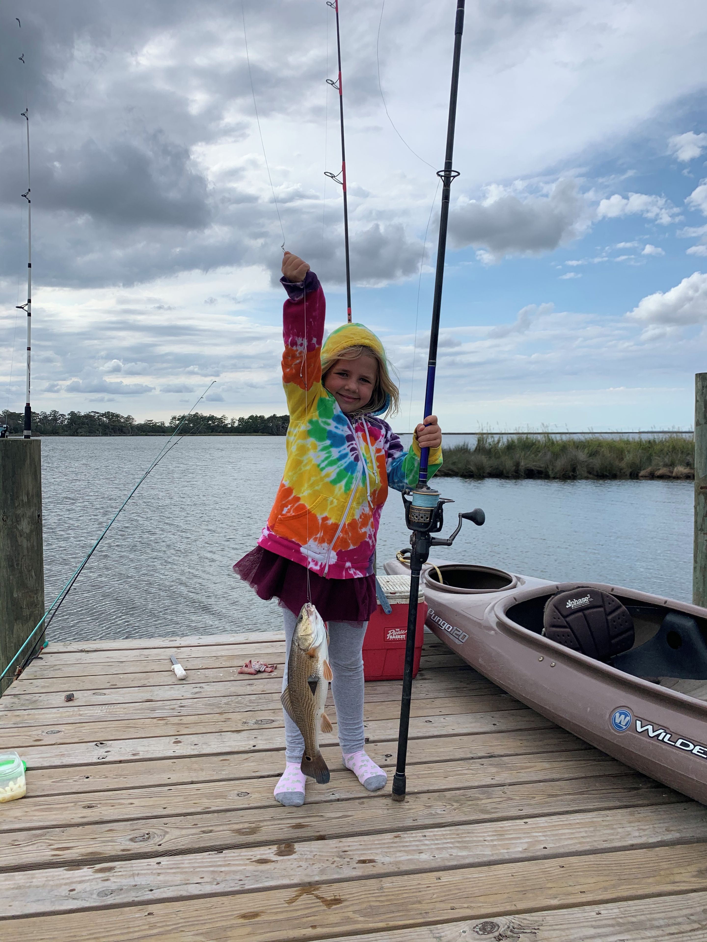 A child on a dock holding a fish, with a canoe and fishing rod in the background.
